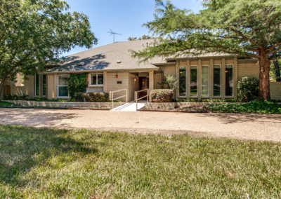 Single-story beige brick house with large vertical windows, a ramp leading to the front door, and surrounded by trees, bushes, and a grassy lawn under a clear blue sky.
