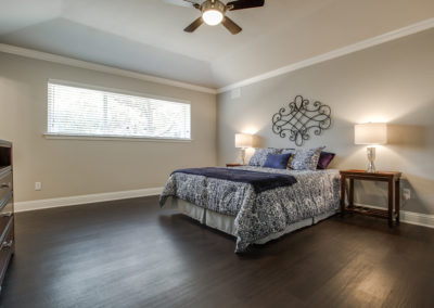 A modern bedroom with dark wood floors, a large bed with blue and white bedding, two bedside tables with lamps, a decorative metal wall piece, and a wide window letting in natural light.