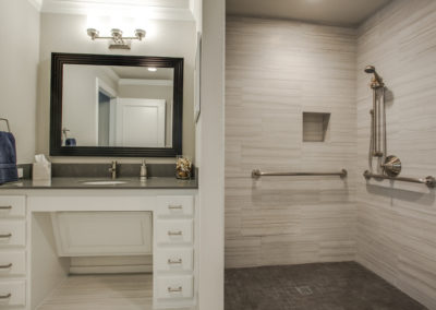 A modern bathroom with a white vanity, a large mirror above a sink, and a walk-in shower with beige tiles, grab bars, a handheld showerhead, and a built-in niche.