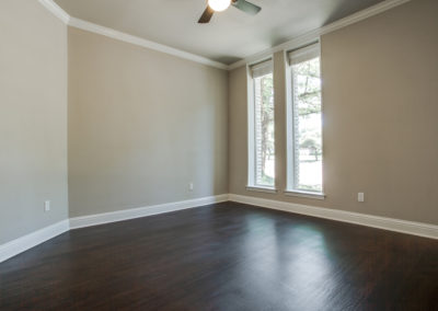 Empty room with light beige walls, dark wood flooring, white baseboards, a ceiling fan, and two tall windows with white blinds, allowing natural light to enter.
