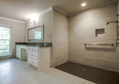 Spacious modern bathroom with a large walk-in shower featuring a bench, grab bar, and handheld showerhead. There’s a long vanity with a mirror and drawers, and a tall window letting in natural light.