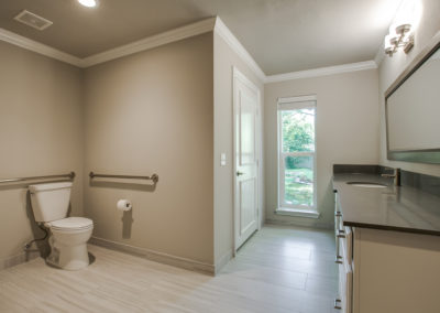 Spacious, modern bathroom with beige walls, a white toilet with grab bars, a long countertop with a sink and cabinets, large mirror, window with a view of greenery, and light wood-look flooring.