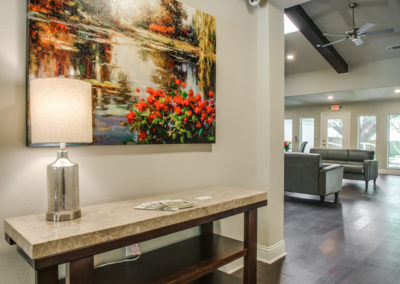 A modern hallway with a wooden console table, lamp, and colorful landscape painting. In the background, there is a seating area with sofas, large windows, and dark wood flooring.