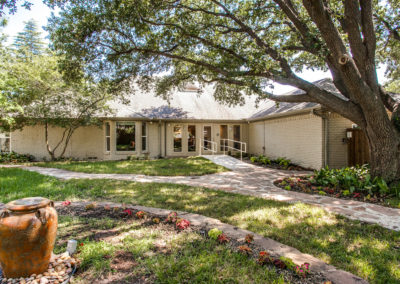 Single-story brick house with large windows, surrounded by lush green grass, trees, and landscaped flower beds. A curved walkway leads to the entrance, which has a ramp for accessibility. An urn sits in the garden in the foreground.