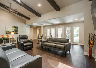 A modern living room with gray leather sofas, a black armchair, flowers on tables, wood floors, ceiling beams, large windows, and a glass door leading to a green outdoor area.