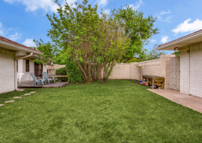 A backyard with green grass, a large leafy tree, a wooden deck with blue chairs, a brick wall, and a workbench along a paved walkway between two white brick buildings under a blue sky.
