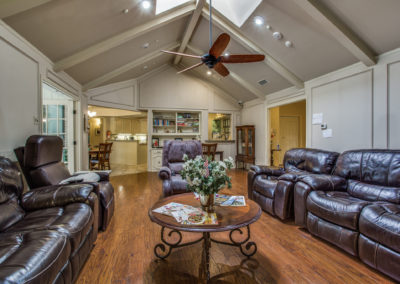 A spacious living room with dark leather recliners, a round coffee table with magazines and flowers, wood flooring, vaulted ceiling with skylights, and a ceiling fan, open to a kitchen and dining area in the background.