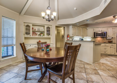A spacious kitchen and dining area with tiled floors, a round wooden dining table with four chairs, a chandelier above, built-in shelves, and light-colored cabinetry with modern appliances in the background.