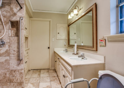 A bathroom with a double sink vanity, large mirror, beige cabinets, tan tiled floor and shower, wall-mounted lights, and a window letting in natural light. A toilet sits near the vanity.