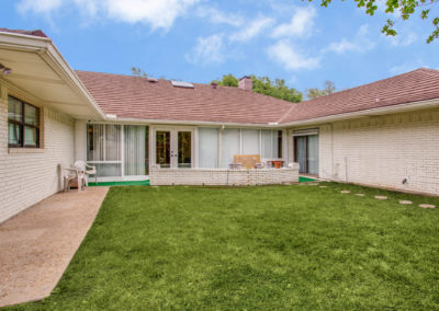 Single-story house with white brick walls and red roof tiles, featuring large windows and sliding glass doors opening to a grassy backyard with a small patio area and outdoor chairs under a partly cloudy sky.