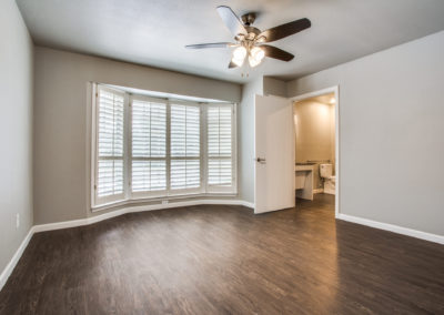 Empty bedroom with wood flooring, large bay window with white shutters, ceiling fan with lights, and an open door leading to a bathroom with visible toilet and vanity. Walls are painted light gray.