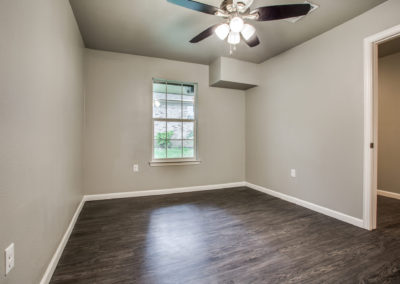 Empty room with gray walls, dark wood flooring, a ceiling fan with lights, and a window letting in natural light. An open doorway leads to another room.