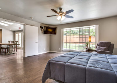 A spacious bedroom with hardwood floors, a ceiling fan, and a large window overlooking a fenced yard. The room includes a bed with a blue comforter, a recliner, and a wall-mounted TV. A dining area is visible through open double doors.