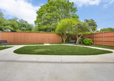 A spacious backyard with a concrete patio, an oval patch of green grass in the center, wooden fencing, benches, and lush trees and shrubs along the fence, under a partly cloudy sky.