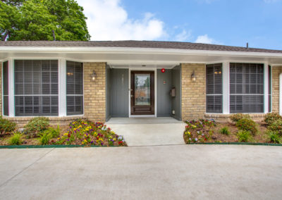 Single-story brick building with large windows, a gray front door, and landscaped flower beds on either side of the entrance under a partly cloudy sky.