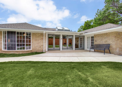 Single-story brick building with large windows, a glass-covered entryway, and a bench beside the entrance. The building is surrounded by green grass and a paved walkway under a partly cloudy sky.
