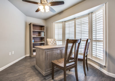 A bright home office with wood flooring, a wooden desk and two chairs, a high-back chair, a bookshelf with a few items, and large windows with white plantation shutters. A ceiling fan with lights is above.