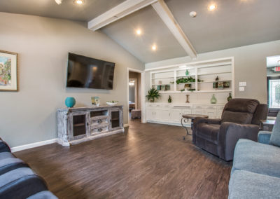 A cozy living room with wood floors, a large wall-mounted TV, plush seating, a rustic TV stand, built-in white shelves with decor, and soft natural lighting.