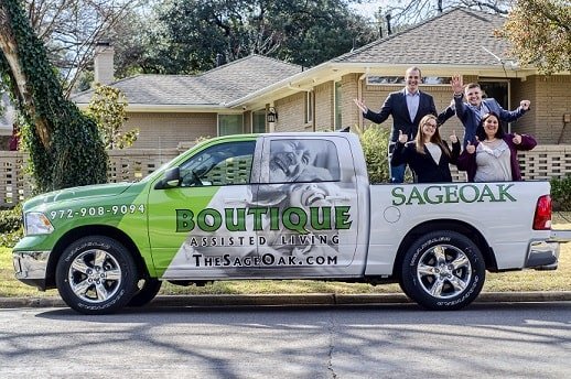 Four smiling people stand in the bed of a green and white pickup truck advertising Sage Oak Boutique Assisted Living, parked in front of a house on a sunny day.