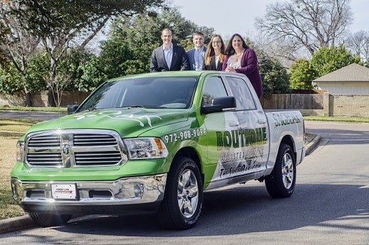 Four people stand in the bed of a green and white pickup truck with Boutique and a phone number printed on the side, parked on a residential street with trees and houses in the background.