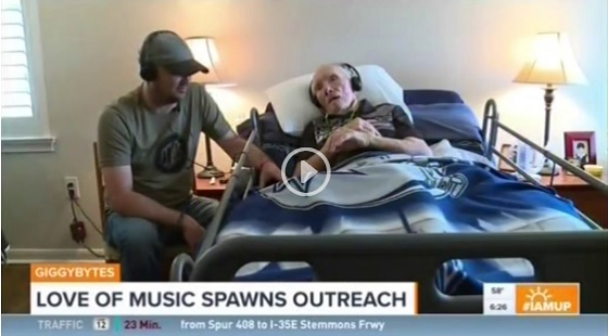 A man wearing headphones sits beside an elderly man in a hospital bed, also wearing headphones. They hold hands, sharing a moment. TV graphics read LOVE OF MUSIC SPAWNS OUTREACH with news and weather info displayed.