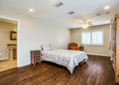 Spacious bedroom with hardwood floors, a bed with gray bedding, a colorful nightstand, a wooden wardrobe, an orange armchair by a window with white shutters, and an en suite bathroom visible to the left.