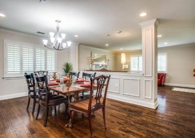 A dining room with a wooden table set for six, featuring red napkins and a chandelier overhead. White paneled walls, hardwood floors, and large windows with shutters brighten the space. A living area is visible in the background.