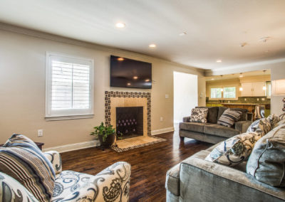 A cozy living room with patterned armchairs, gray sofas, a fireplace with tile surround, a wall-mounted TV above the mantel, and a potted plant, all facing hardwood floors and an open view into the kitchen.