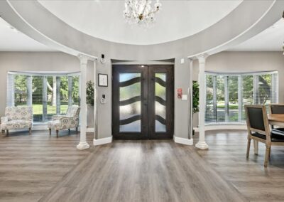 Bright, elegant foyer with large double doors, glass panels, columns, and a chandelier. Two armchairs and a dining table sit by curved windows with blinds, showcasing a view of green trees outside.