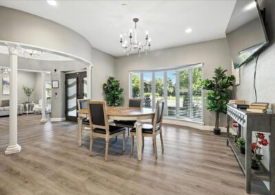 A modern dining room with a round wooden table and four black chairs, large windows with a view of greenery, light wood flooring, a chandelier, and decorative plants and shelves.