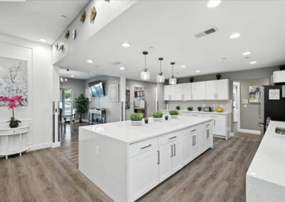 Modern kitchen with white cabinets and countertops, large island with potted plants, pendant lights, stainless steel appliances, and wood flooring. Open layout leads to a dining area and living room in the background.