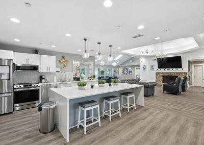 Modern open-concept kitchen and living area with white cabinets, stainless steel appliances, a large island with stools, pendant lighting, and a cozy living room featuring a fireplace and sectional sofa.