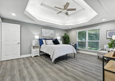 A modern bedroom with gray walls, light wood floors, a large bed with blue and white bedding, lamps on white nightstands, a potted plant, a ceiling fan, and a large window with blinds.