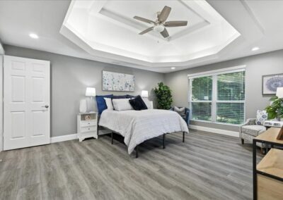 Spacious bedroom with gray walls, wood floors, a large bed with white and blue bedding, nightstands, lamps, a potted plant, two chairs by a large window, and a tray ceiling with a ceiling fan.