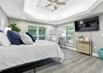 Modern bedroom with gray walls, wood floors, a large bed with blue and white pillows, a wall-mounted TV, ceiling fan, armchair, plants, and large windows letting in natural light.