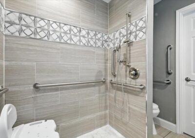Modern accessible shower with beige tile, decorative accent strip, multiple stainless steel grab bars, handheld showerhead, shower chair, and an open entry leading to a bathroom with a white door and additional grab bar.