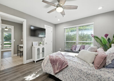A bright, modern bedroom with a ceiling fan, wall-mounted TV, and a bed with pastel bedding and pillows. Large window with blinds lets in sunlight, and there is a leafy plant and a white cabinet in the room.