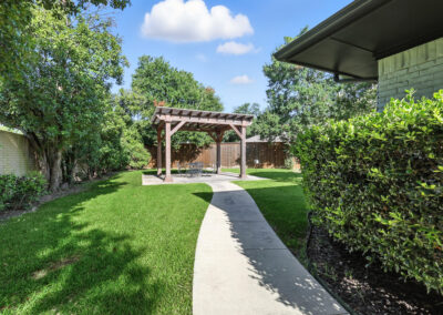 A concrete path curves through a green backyard toward a wooden pergola with chairs underneath, surrounded by trees, bushes, and a wooden fence on a sunny day.